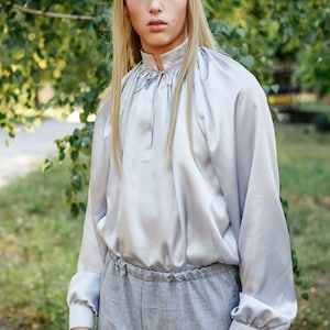 May include: A person with long blonde hair wearing a light grey satin shirt with a gathered neckline and long sleeves. They are also wearing grey trousers with a drawstring waist.