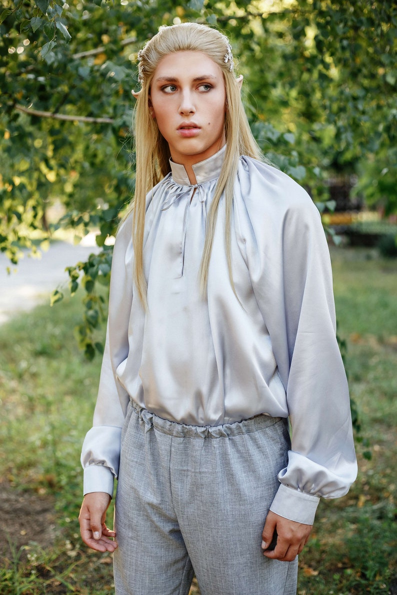May include: A person with long blonde hair wearing a light grey satin shirt with a gathered neckline and long sleeves. They are also wearing grey trousers with a drawstring waist.