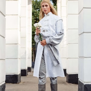 May include: A person wearing a white long-sleeved tunic with a silver sash and grey trousers. They are holding a bouquet of white flowers and wearing grey boots.