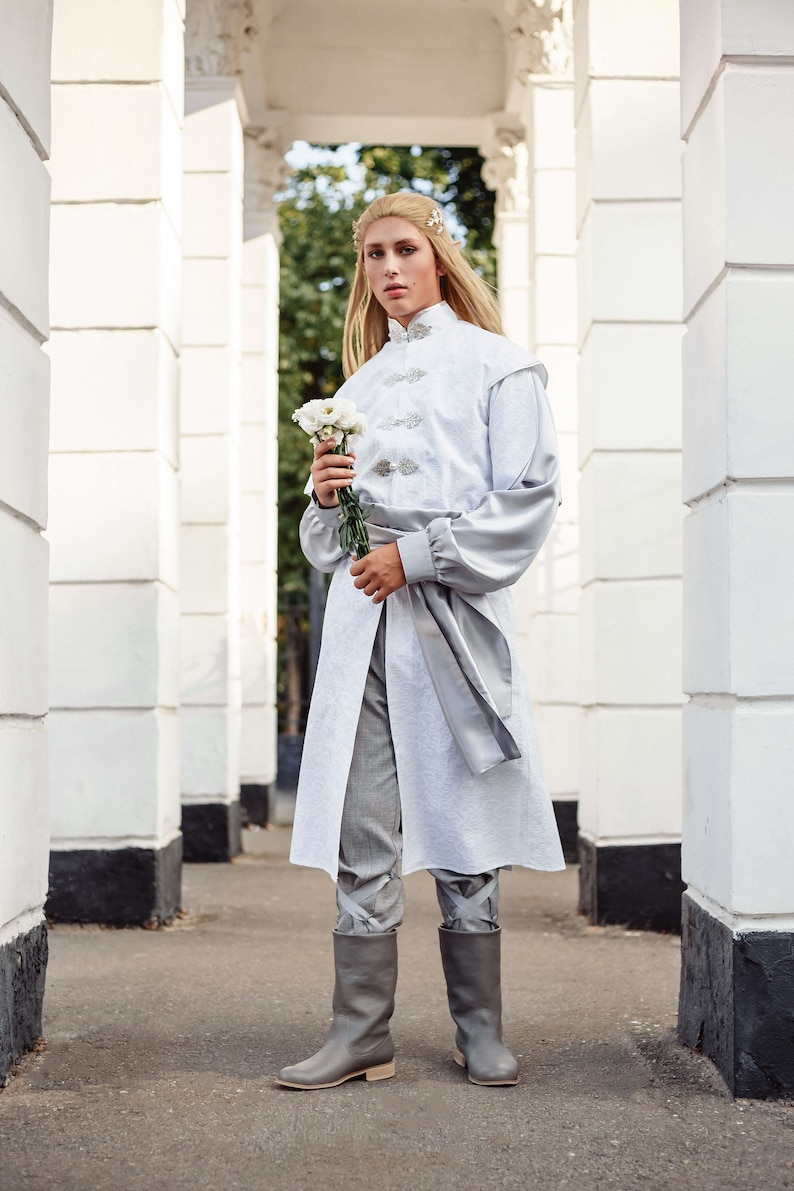 May include: A person wearing a white long-sleeved tunic with a silver sash and grey trousers. They are holding a bouquet of white flowers and wearing grey boots.