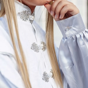 May include: A white blouse with a high collar and pearl buttons. The blouse features intricate silver embroidery on the collar and cuffs. The sleeves are long and puffy.