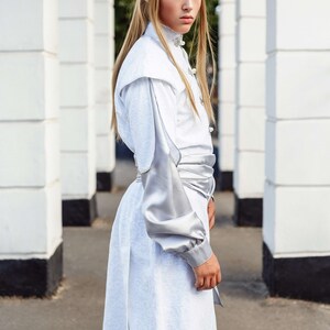 May include: A person wearing a white and silver costume with long blonde hair and pointed ears. The costume has a silver sash and a silver trim on the sleeves. The person is standing in front of a white wall.