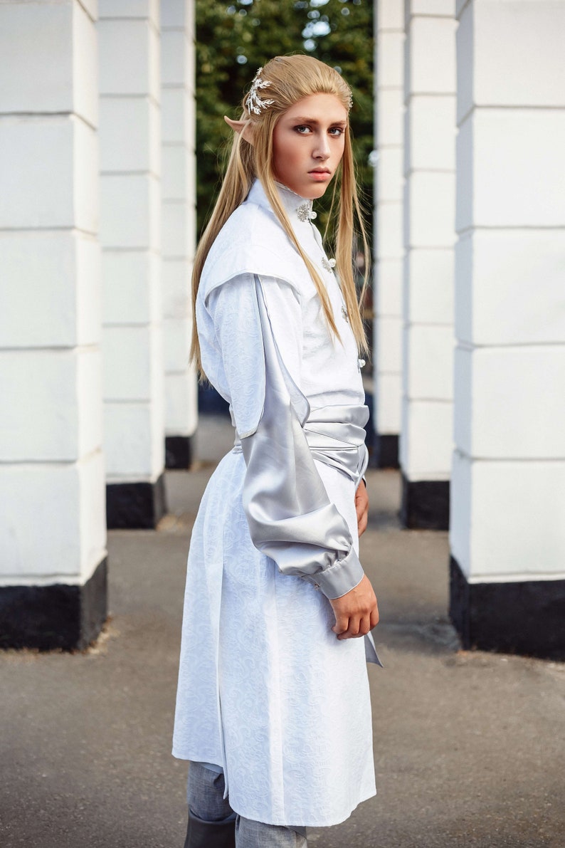 May include: A person wearing a white and silver costume with long blonde hair and pointed ears. The costume has a silver sash and a silver trim on the sleeves. The person is standing in front of a white wall.