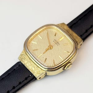 May include: A gold-toned Seiko Quartz watch with a square face and a black leather band. The watch face has a textured pattern and gold hands. The watch case is gold-colored with a textured finish. The watch band is black.