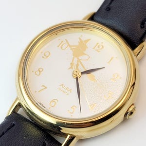May include: Gold-tone Alba Quartz watch with a white face featuring a witch design. The watch has gold numerals, black hands, and a black leather strap. The watch is a classic, elegant accessory.