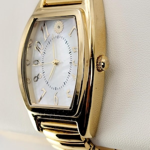May include: A gold wristwatch with a rectangular face and a white mother-of-pearl dial. The watch features gold numerals and hands, and a gold band. The watch is displayed on a white surface.