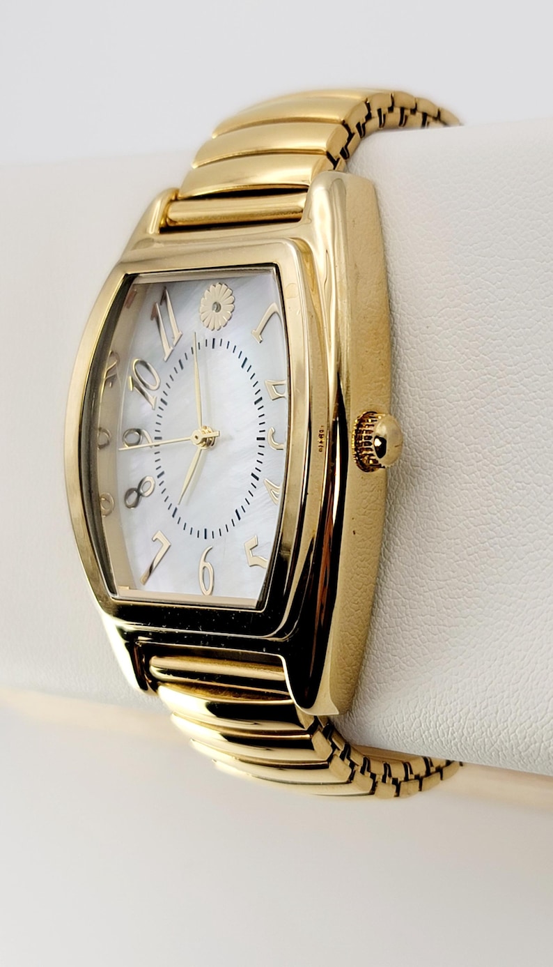 May include: A gold wristwatch with a rectangular face and a white mother-of-pearl dial. The watch features gold numerals and hands, and a gold band. The watch is displayed on a white surface.