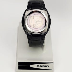 Op de afbeelding: Zwarte Baby-G digitale horloge met een vierkante wijzerplaat en zwarte band. De wijzerplaat toont de tijd en het woord "NYC". Het horloge staat op een witte displaystandaard met het Casio-logo.