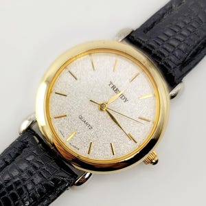 Op de afbeelding: Een goudkleurig quartz horloge met een zilveren wijzerplaat en een zwarte leren band. De wijzerplaat heeft gouden wijzers en markeringen, met het woord "TRENDY" boven het midden gedrukt. De band is ongeveer 2 cm breed.