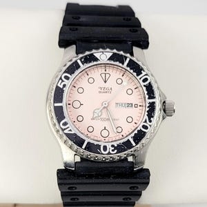May include: A silver-toned VEGA Quartz watch with a black bezel and band. The watch face is light pink with black markings and the date is displayed. The watch is water resistant to 100 meters.