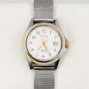 May include: A silver-toned wristwatch with a gold-colored bezel and a white face. The watch has a mesh band and displays the time with black numerals and hands. The word "Cassy" is printed on the watch face.