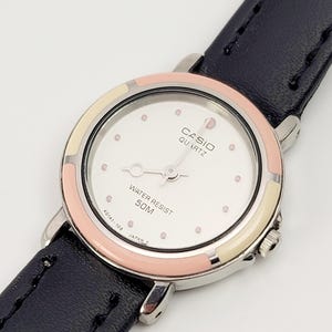 May include: A Casio Quartz watch with a white face, pink and cream-colored bezel, and a black leather band. The watch face has pink dot hour markers and the words "CASIO QUARTZ" and "WATER RESIST 50M".