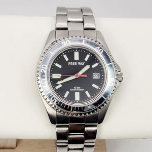 May include: A stainless steel analog wristwatch with a black dial, featuring the text FREE WAY and 10 bar WATER RESIST. The watch has a rotating bezel with numerical markers, luminous hour markers, a date window at the three o'clock position.