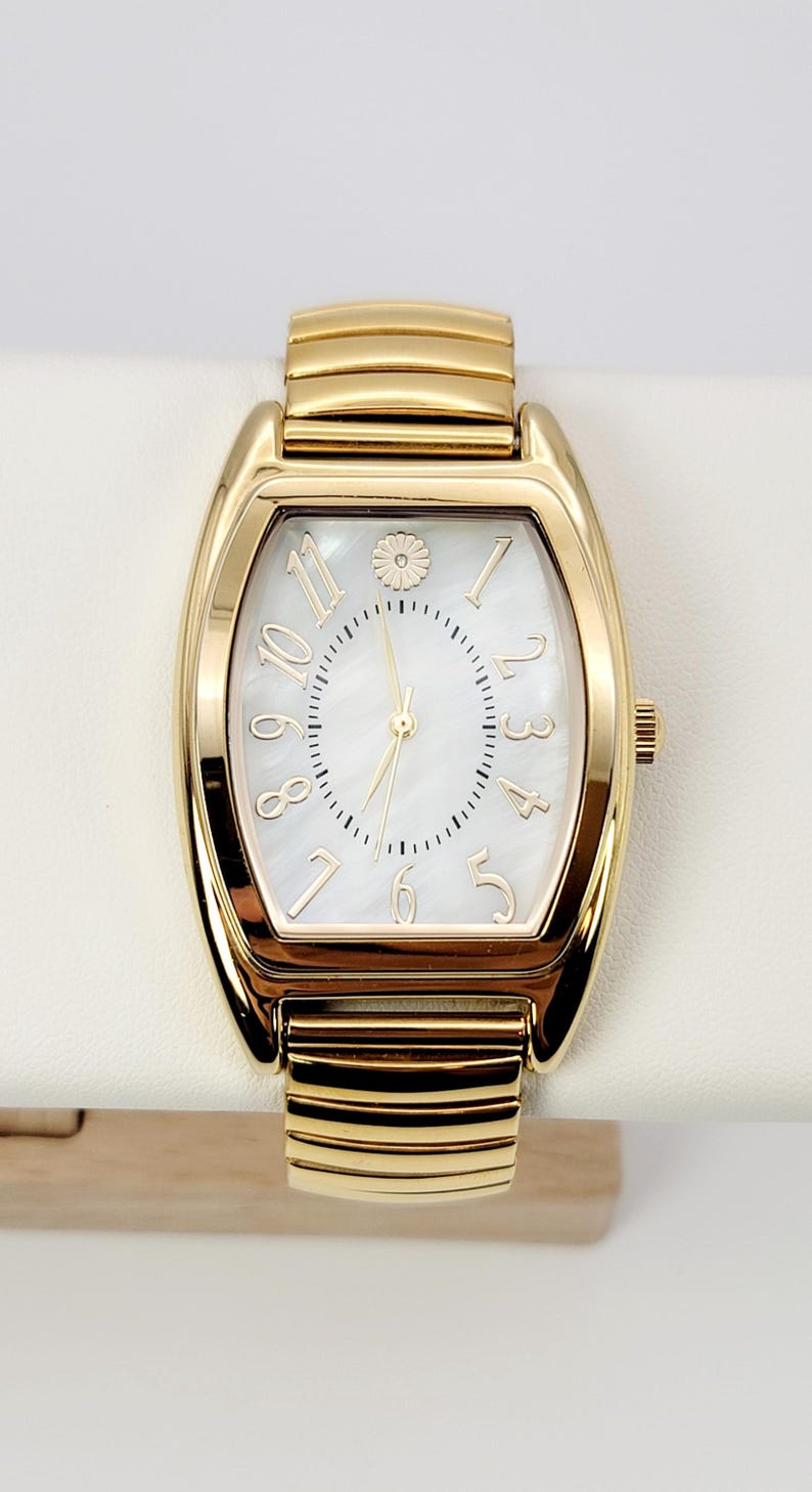May include: Gold wristwatch with a rectangular face and a white mother-of-pearl dial. The watch features gold hands and hour markers, with a small floral detail at the 12 o'clock position. The watch band is gold-toned with a ribbed design.