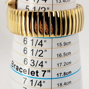 May include: A gold-coloured bracelet, displayed on a white measuring tool. The bracelet is approximately 7 inches (17.8 cm) in circumference. The measuring tool has black text and blue lines indicating bracelet sizes in inches and centimetres.