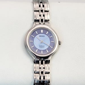 Puede incluir: Un reloj de pulsera Seiko Exceline Titanium plateado con esfera azul claro. El reloj tiene una correa de metal con eslabones y una esfera redonda. Se ven el nombre de la marca y los detalles del modelo. El reloj está sobre una superficie blanca.