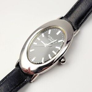 Könnte beinhalten: Silberfarbene ovale Uhr mit schwarzem Zifferblatt und schwarzem Lederarmband. Das Zifferblatt hat silberne Stundenmarkierungen und Zeiger mit dem Text "DURALAY LONDON". Die Uhr hat eine kleine, runde Krone.