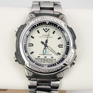 May include: Silver-tone Casio watch with a white face, featuring black hands and markers. The watch has a digital display showing "1:3426" and the word "ILLUMINATOR" is visible on the bezel. The watchband is made of silver metal.