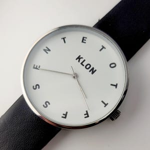 Puede incluir: Un reloj de pulsera minimalista con esfera blanca y manecillas negras. La esfera del reloj presenta la palabra "KLON" y números negros. El reloj tiene un bisel plateado y una correa de cuero negro.