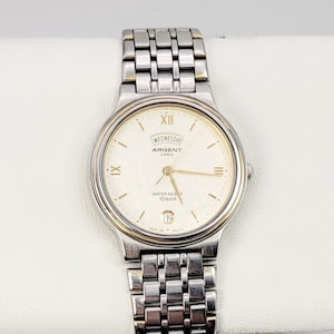May include: Silver-toned Argent watch with a gold-colored bezel and a silver link band. The watch face is cream-colored with Roman numerals, the day of the week, and the date. The watch is water resistant to 10 bar.