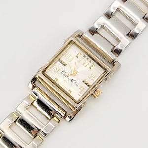 May include: A rectangular wristwatch with a silver and gold-tone metal strap. The watch face is white with gold hands and numerals. The brand name "Cecil McBee" is printed on the watch face.