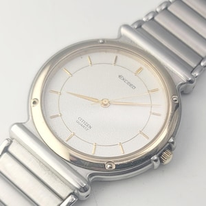 May include: A Citizen Exceed quartz wristwatch featuring a round white textured dial with gold-tone hour markers and hands. The watch has a two-tone silver and gold bezel and a silver-tone metal link bracelet with a polished finish.
