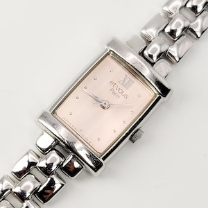 May include: A rectangular wristwatch with a pale pink face and a silver-toned metal strap. The watch face features Roman numerals and the words "et vous Paris". The strap is made of connected rectangular links.