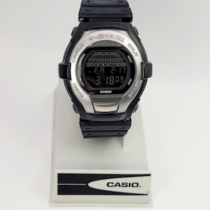 Op de afbeelding: Zwarte en zilveren G-Shock digitale horloge met een rechthoekig display met datum en tijd. Het horloge heeft meerdere knoppen en de merknaam "CASIO" is zichtbaar op de wijzerplaat en de standaard.