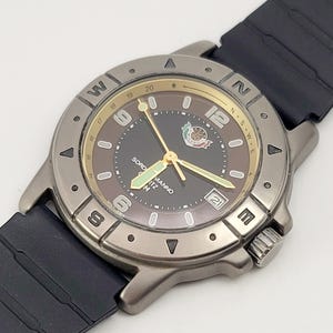 May include: A wristwatch with a dark brown face, gold hands, and a date display. The watch has a silver-tone bezel with compass markings and a black rubber strap. The watch face has the brand name "Sord Manno" printed on it. The watch is a stylish accessory.