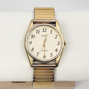 May include: Gold-toned Seiko Quartz Chronos watch with a cream-colored face and gold band. The watch features black numerals and hands, with the brand name and model name visible on the face. The band is a flexible, expandable style.