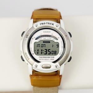 Op de afbeelding: Een Casio Pro Trek digitale horloge met een witte wijzerplaat, zilveren rand en bruine band. Het horloge geeft de tijd, datum en andere functies weer. Het horloge heeft een thermometer en is waterdicht tot 10 bar.