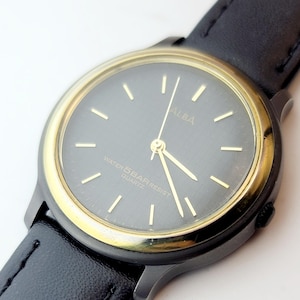 May include: A black ALBA quartz watch with a gold-colored bezel and a black leather band. The watch face is dark gray with gold-colored hands and markers. The watch is marked "WATER 5 BAR RESIST QUARTZ".