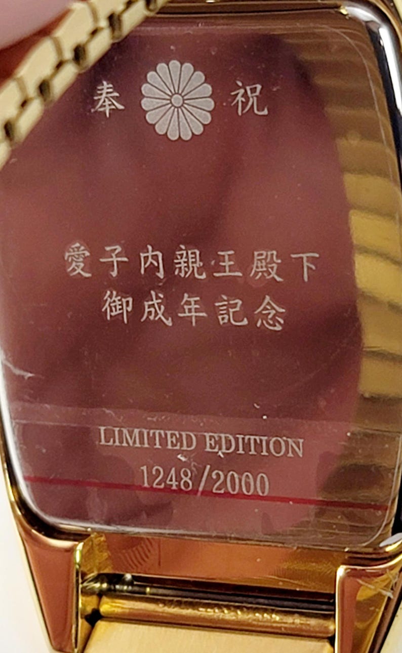 May include: A gold watch with a burgundy face, featuring Japanese characters and a white floral emblem. The text "LIMITED EDITION 1248/2000" is displayed on the watch face. A limited edition timepiece.