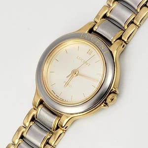 May include: A gold and silver-toned Lucent watch with a cream-colored face. The watch features gold hands and hour markers, with the word "LUCENT" printed on the face. The watch band is a combination of gold and silver links.