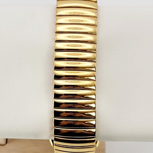 May include: Close-up of a gold-toned watch strap. The strap is composed of rectangular links, each with a slightly curved surface, creating a textured appearance. The watch strap is displayed on a white surface.