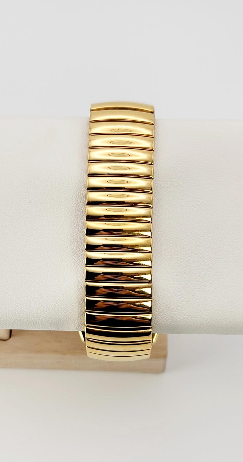May include: Close-up of a gold-toned watch strap. The strap is composed of rectangular links, each with a slightly curved surface, creating a textured appearance. The watch strap is displayed on a white surface.