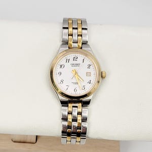 May include: A gold and silver-toned Orient Quartz wristwatch with a white face. The watch features gold hour markers and hands, a date window at the 3 o'clock position, and a silver and gold link band. The watch is approximately 1.25 inches in diameter.