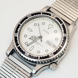 May include: Silver-toned Alba quartz watch with a white face, black bezel, and a date display showing "THU 15." The watch has a silver metal band. The watch face features geometric hour markers and the text "5BAR RESIST QUARTZ."