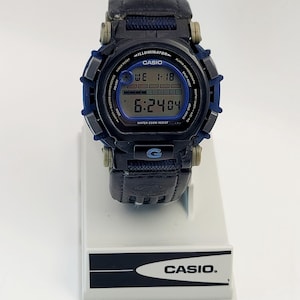 Peut inclure: Montre numérique Casio noire et bleue, affichant la date et l'heure. La montre est dotée d'un bracelet en cuir noir et est résistante à l'eau. Elle est présentée sur un présentoir blanc avec le logo Casio.