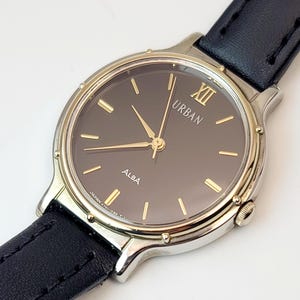 May include: A silver-toned ALBA URBAN watch with a dark brown face and gold-colored hands and markers. The watch has a black leather band. The watch face displays the Roman numeral XII.