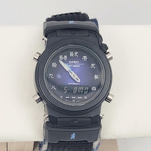 May include: A black Casio G-Shock watch with a dark blue analog dial featuring Japanese kanji numerals. The watch includes a digital display at the bottom, silver-toned hands, and a black fabric strap with blue accents and a kanji character on the lug.