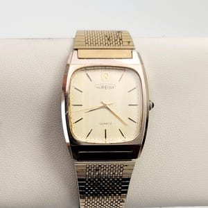 May include: Aureole quartz watch with a rectangular gold-toned face and a silver and gold-toned band. The watch face is cream-colored with black markers and hands. The watch band has a textured design.