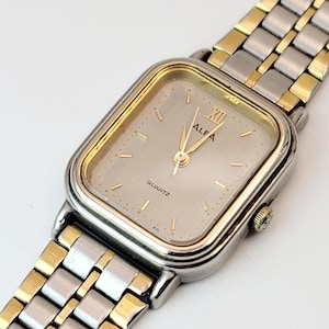 May include: A rectangular ALFA quartz watch with a silver and gold-tone band. The watch face is a light silver color with gold-tone hands and markers. The watch band is a combination of silver and gold-tone links.