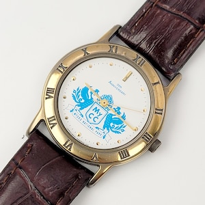 Puede incluir: Un reloj de pulsera dorado con esfera blanca y correa de cuero marrón. La esfera del reloj presenta números romanos, detalles dorados y un emblema azul con el texto "My CC" y "Myoko National Park". La correa es de cuero marrón oscuro texturizado.