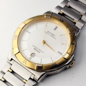 May include: A Sting Sporty Version wristwatch with a silver and gold-tone metal band and bezel. The watch face is white with gold hands and Roman numeral hour markers. The watch also features a date display at the 3 o'clock position.