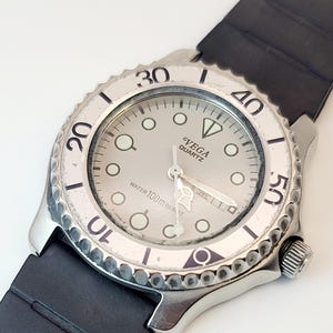 May include: A silver-toned Vega Quartz watch with a black rubber strap. The watch face is light gray with black numerals and luminous markers. The bezel has a textured edge and the watch is marked "Water 100m Resist".