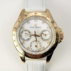 Könnte beinhalten: Eine goldfarbene Armbanduhr mit weißem Zifferblatt und weißem Lederarmband. Das Zifferblatt der Uhr trägt den Text "DOLCE SEGRETO" und mehrere Hilfszifferblätter. Die Uhr hat eine goldene Lünette mit schwarzen Markierungen und Diamantakzenten.