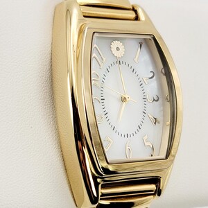 May include: Gold-coloured rectangular wristwatch with a white face and gold numerals. The watch features a gold band and a small floral design at the 12 o'clock position. The watch is displayed on a white surface.