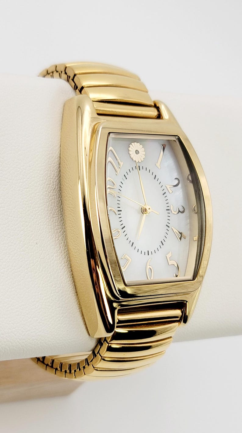 May include: Gold-coloured rectangular wristwatch with a white face and gold numerals. The watch features a gold band and a small floral design at the 12 o'clock position. The watch is displayed on a white surface.