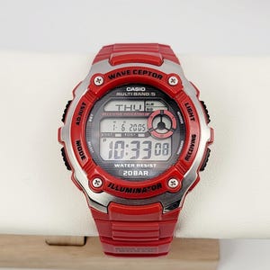 Op de afbeelding: Rood Casio Wave Ceptor digitale horloge met een zilveren rand. De wijzerplaat toont de tijd, datum en dag. Waterbestendig tot 20 bar. Het horloge heeft meerdere knoppen om instellingen aan te passen.